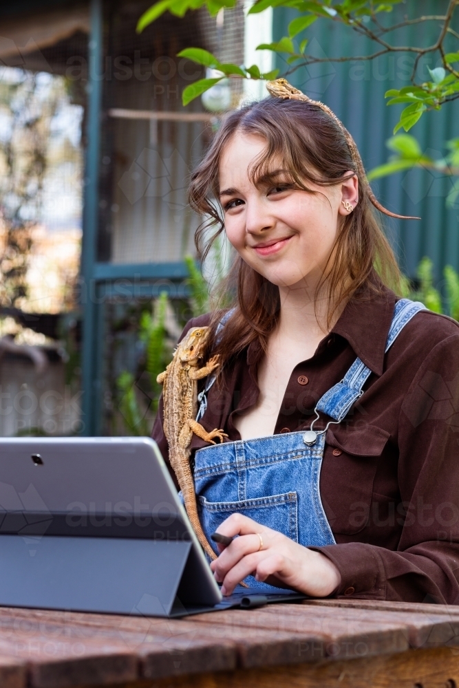Happy young woman sitting at picnic bench in garden with pet lizards working on tablet laptop - Australian Stock Image