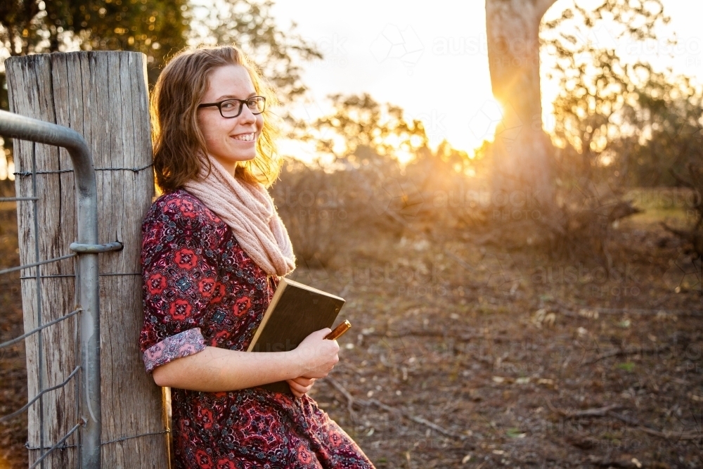 Happy young writer holding pen and notebook leaning on fence post in paddock - Australian Stock Image
