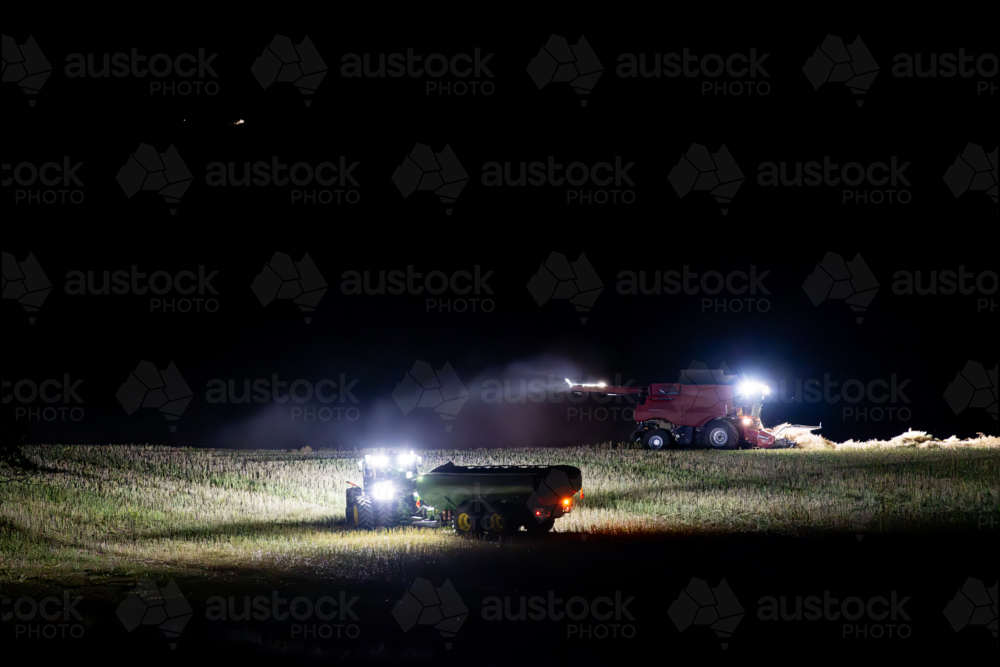 Harvester harvesting canola crop at night with chaser bin on farm - Australian Stock Image
