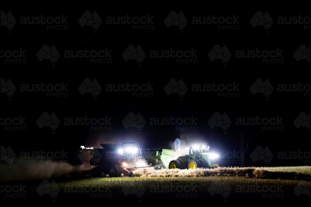 Harvester harvesting canola crop at night with chaser bin on farm - Australian Stock Image
