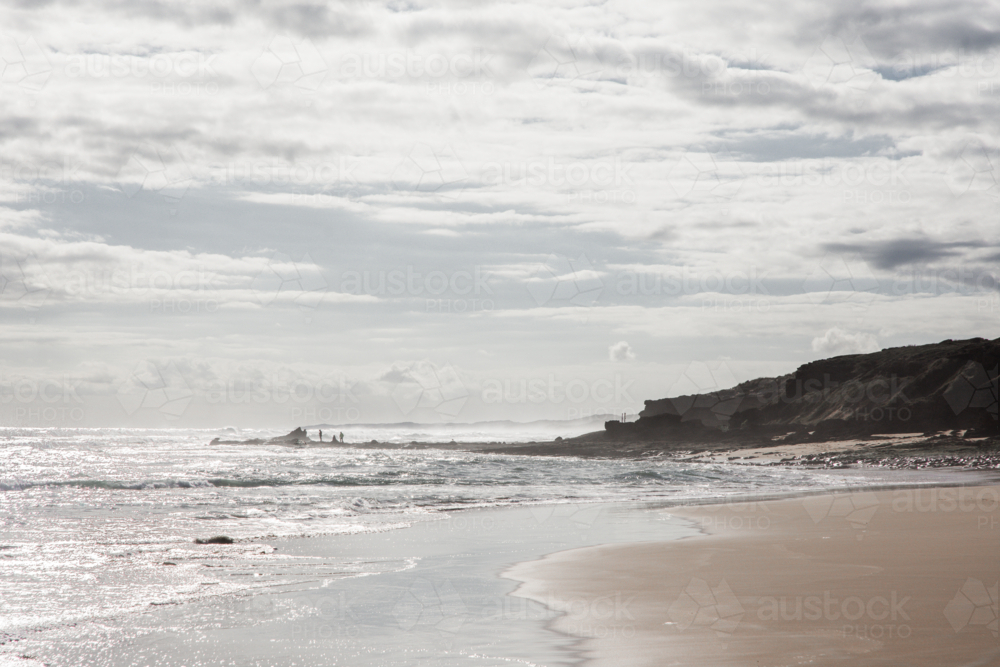 Hazy, late afternoon beach with people on rocks in the distance - Australian Stock Image