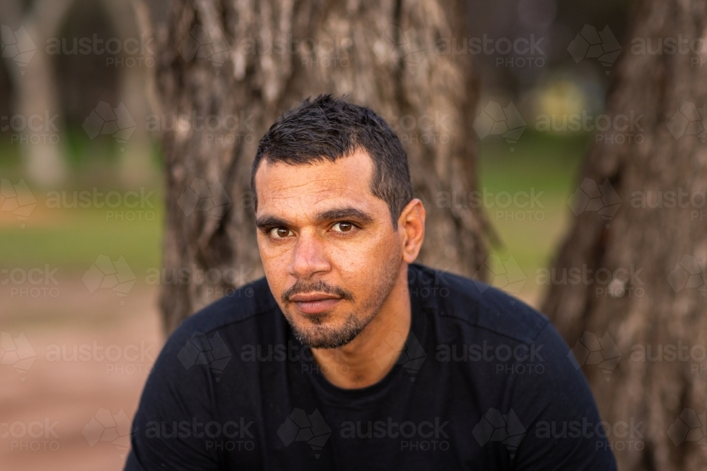 head and shoulders of aboriginal man in black tee shirt sitting in front of a tree - Australian Stock Image
