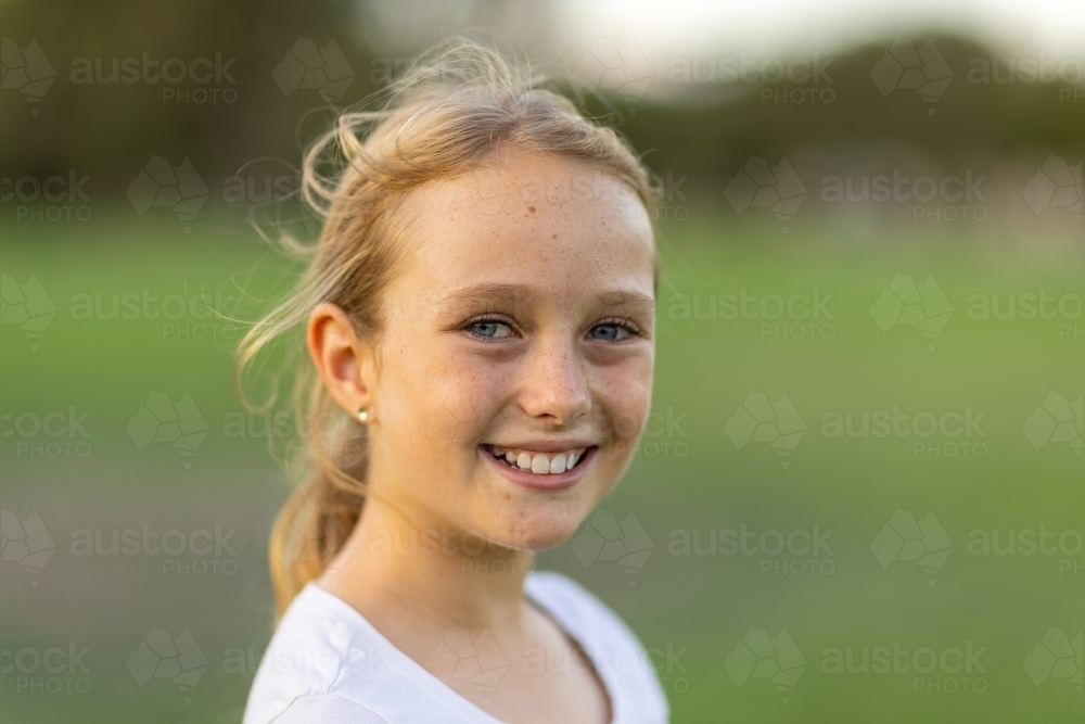 head and shoulders of blonde ten-year-old girl smiling and looking at camera with blurry background - Australian Stock Image