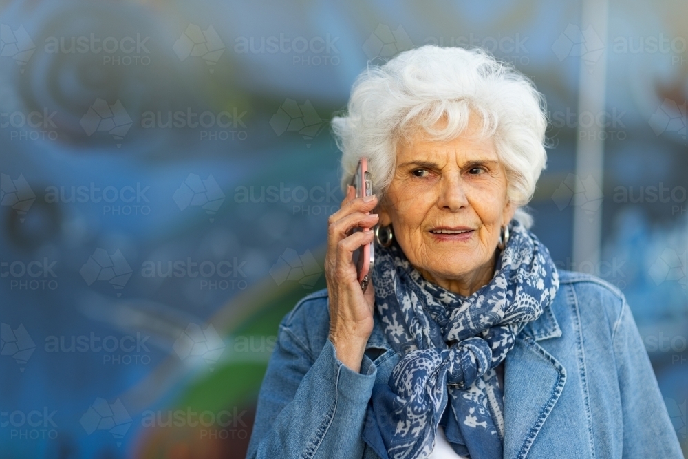 head and shoulders of white-haired elderly lady holding phone to ear with blurred background - Australian Stock Image