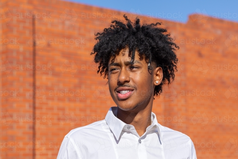 head and shoulders of young man squinting into sun in front of red brick wall - Australian Stock Image