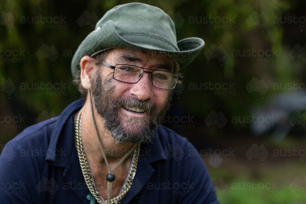 Head and shoulders portrait of a homeless man wearing a hat and glasses, and smiling - Australian Stock Image
