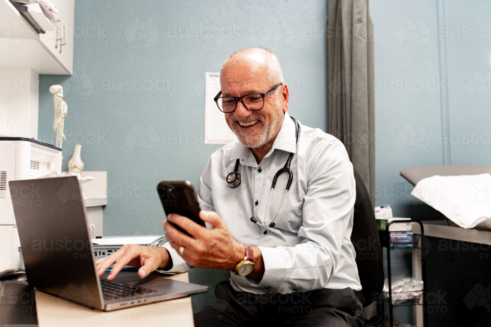 Healthcare professional at desk smiling while working on his laptop and checking smartphone - Australian Stock Image
