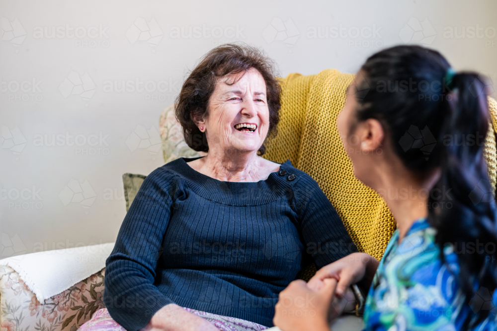 Healthcare worker engaged in a conversation with an elderly woman seated on patterned armchair - Australian Stock Image