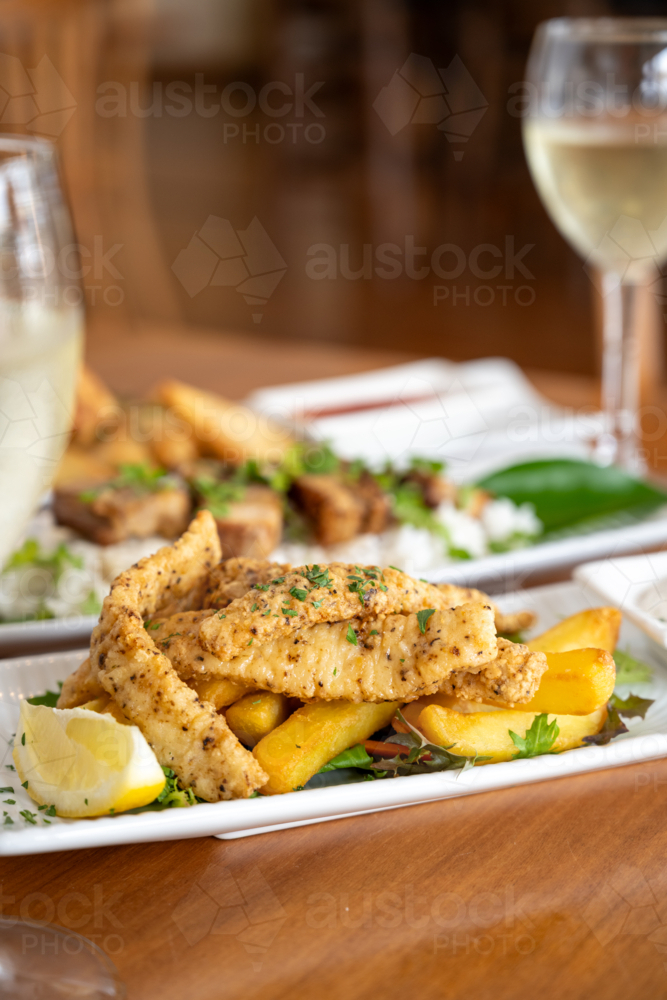 Herb-seasoned fish fillets alongside crispy potato wedges and fresh lemon wedges on white plate - Australian Stock Image