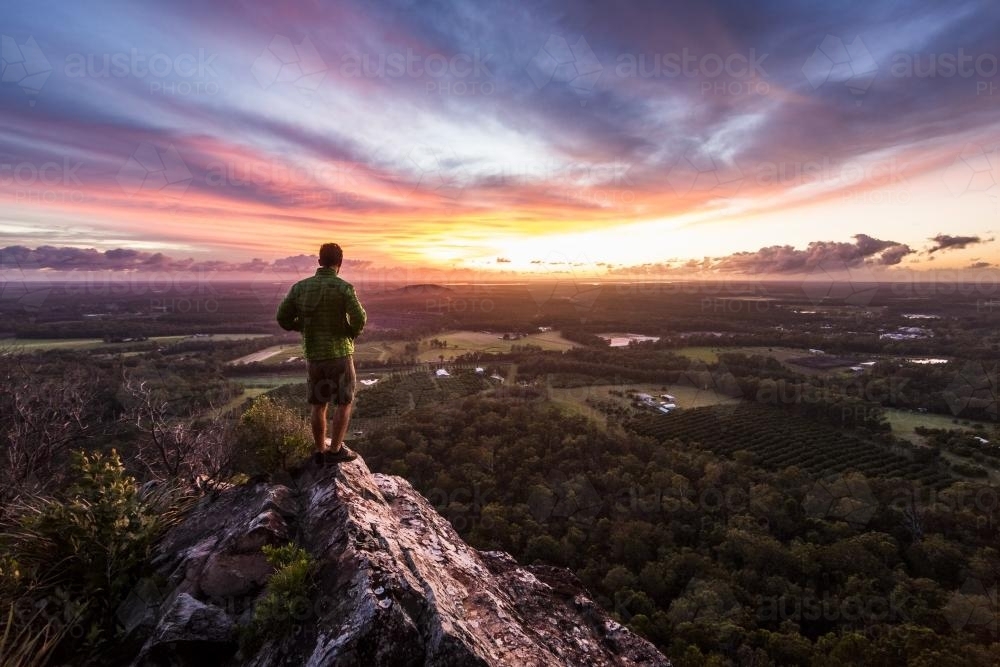 Hiker standing atop a rock formation watching sunrise over valley - Australian Stock Image