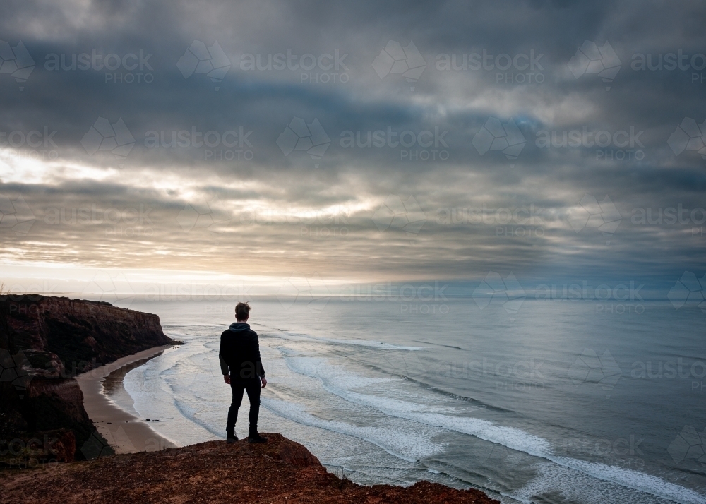 Hiker Standing on a Great Ocean Road Cliff Overlooking a Moody Beach - Australian Stock Image
