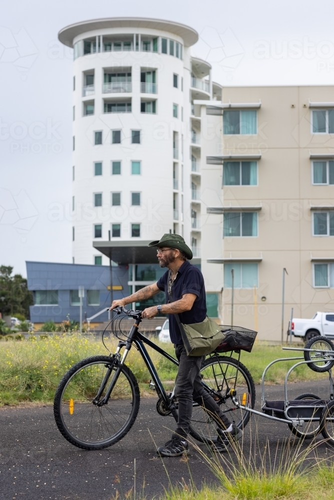 homeless man pushing bike with trailer in front of silo apartments - Australian Stock Image