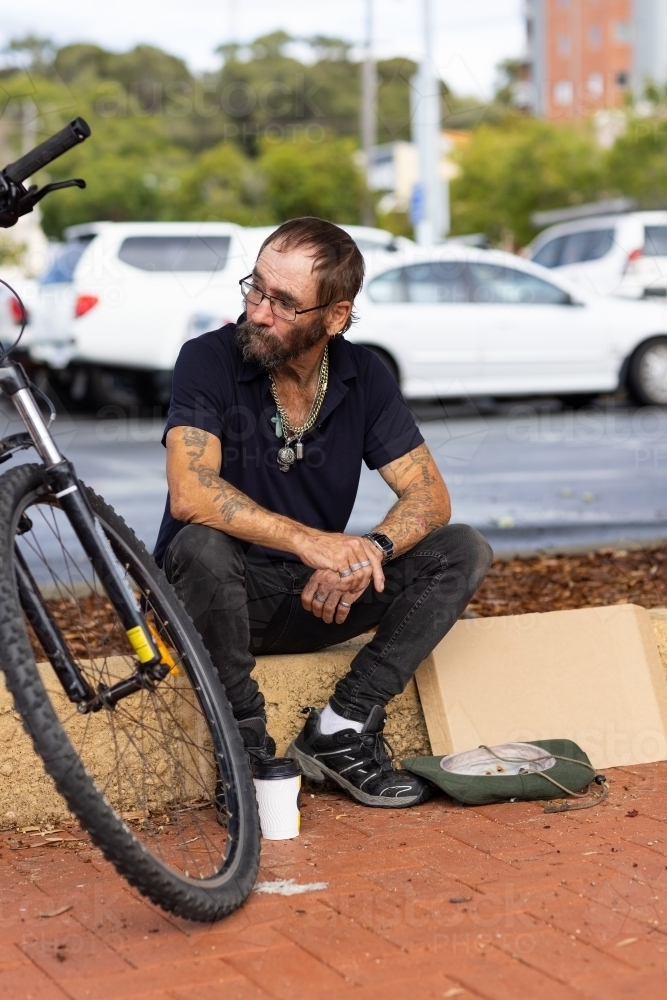 homeless man sitting beside footpath near parking area - Australian Stock Image