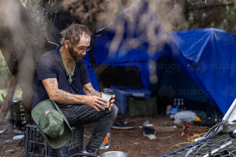 Homeless man sitting on crate outside his tent with a coffee - Australian Stock Image