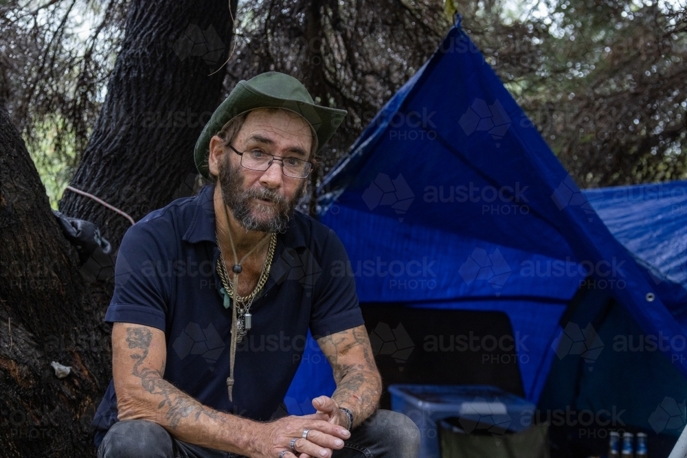 homeless man sitting under trees where he has set up a tent for shelter - Australian Stock Image
