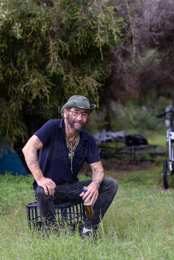 homeless man with beer sitting on milk crate and camp behind him - Australian Stock Image