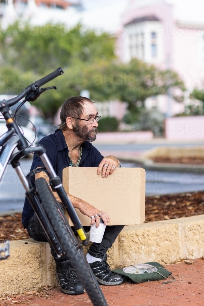 homeless man with bicycle sitting on low wall with cardboard sign - Australian Stock Image