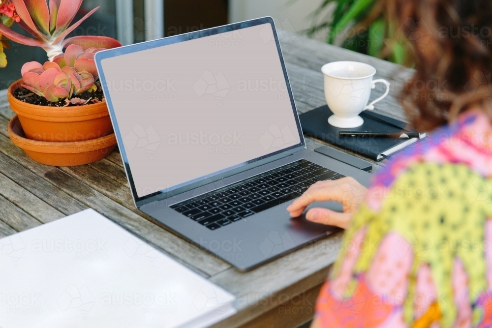 horizontal pic of a person using a laptop on a table with white tea cup and plants on a pot - Australian Stock Image