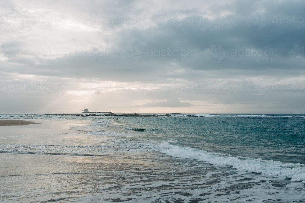 horizontal shot of a beach with waves, rocks and ship at a distance on a cloudy day - Australian Stock Image