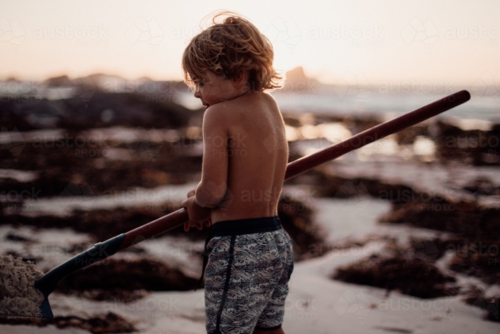 Horizontal shot of a boy holding a shovel with sand by the beach - Australian Stock Image