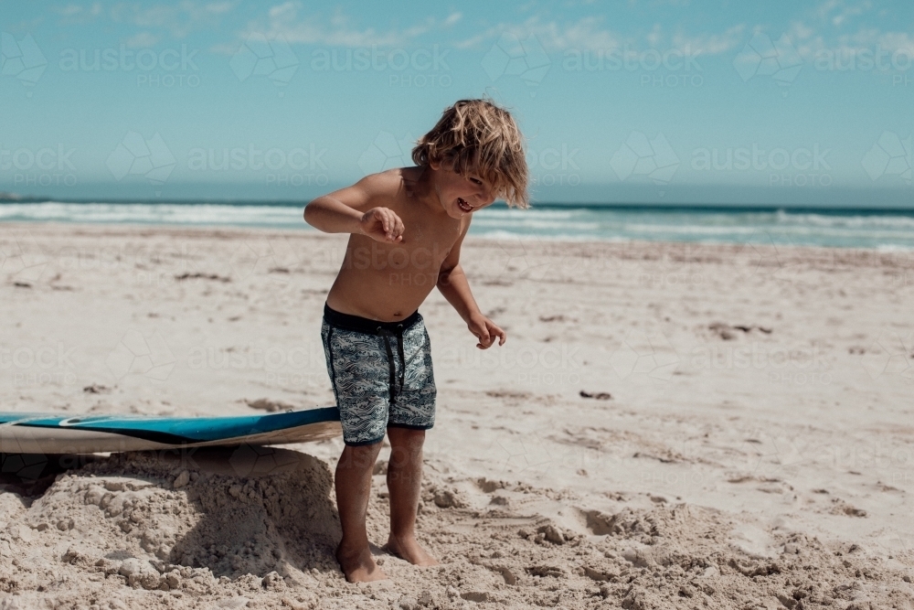 Horizontal shot of a boy playing on the sand beach with a surf board behind him. - Australian Stock Image