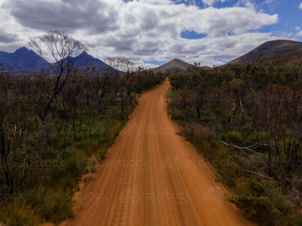 horizontal shot of a dirt road surrounded by trees, grass and bushes with mountains and cloudy skies - Australian Stock Image