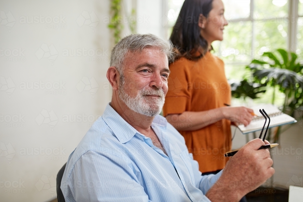 Horizontal shot of a man sitting and smiling while holding a pen and his eye glasses - Australian Stock Image