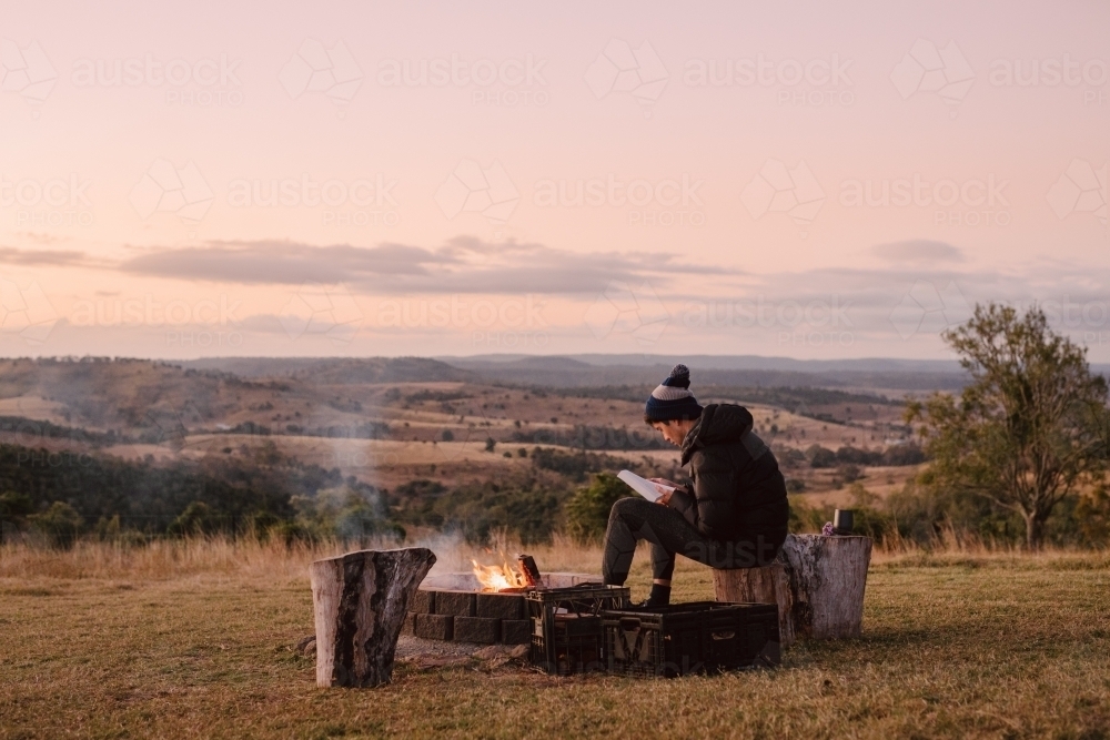 Horizontal shot of a man sitting on a log near a camp fire on a hill top while reading a book - Australian Stock Image