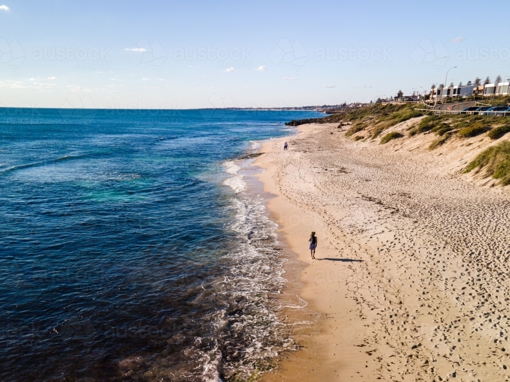 horizontal shot of a person walking on the beach with bushes and small waves on a sunny day - Australian Stock Image