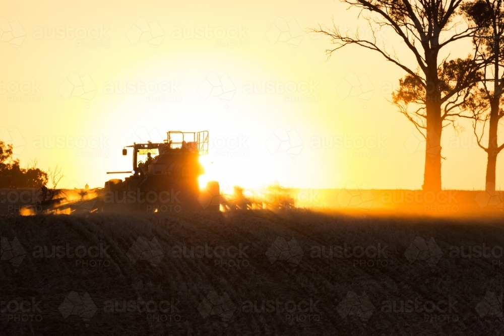 horizontal shot of a silhouette of a tractor, shrubs and two trees with a sunset in the background - Australian Stock Image