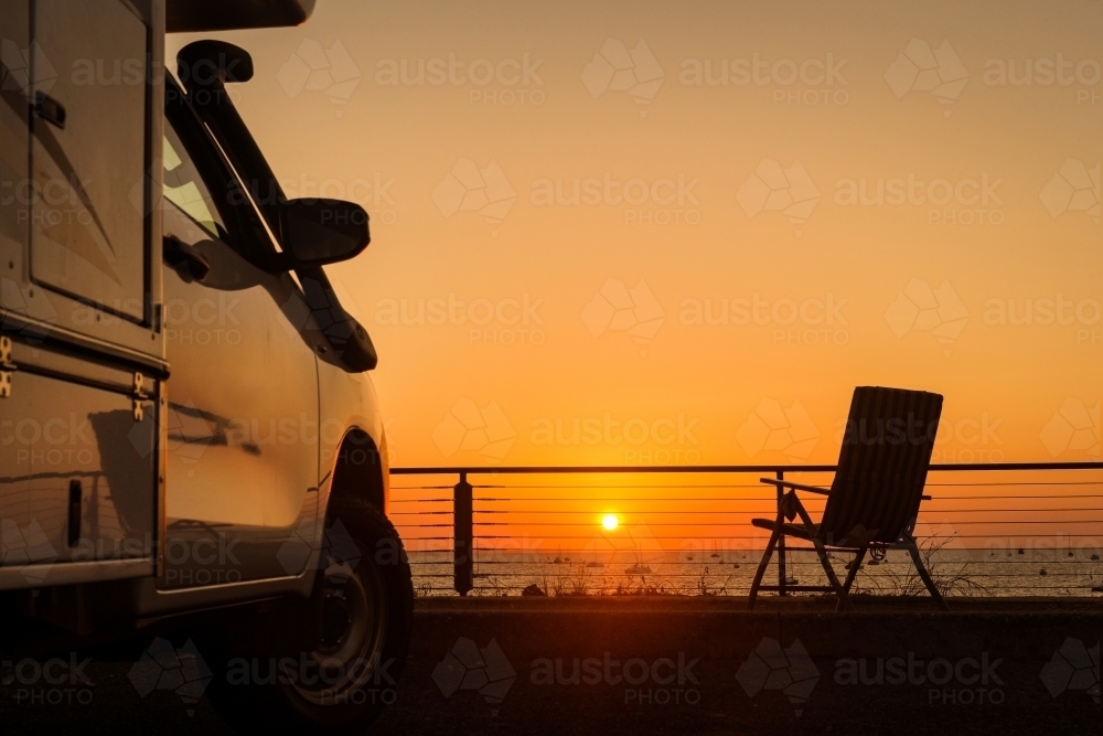 Horizontal shot of a white camper van at sunset in front of the sea with a foldable chair - Australian Stock Image