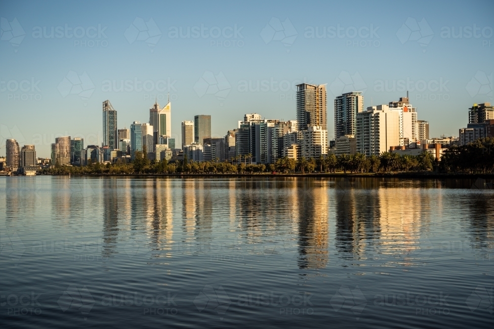 horizontal shot of buildings with trees and reflections appearing in a lake under clear blue skies - Australian Stock Image