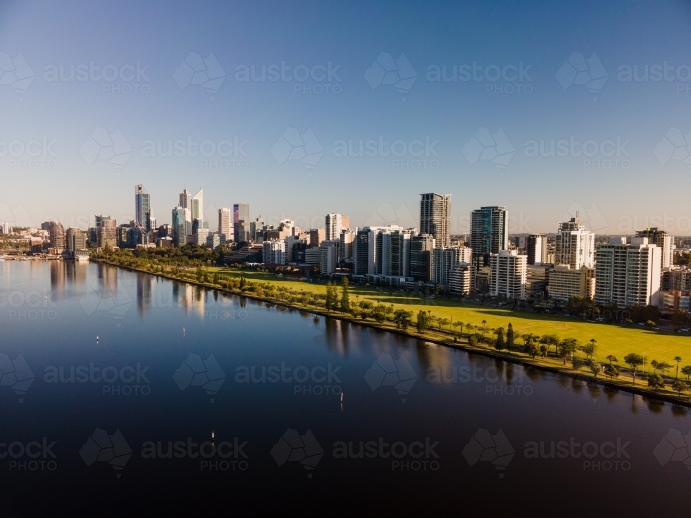 horizontal shot of buildings with trees reflecting in a lake under clear blue and white skies - Australian Stock Image