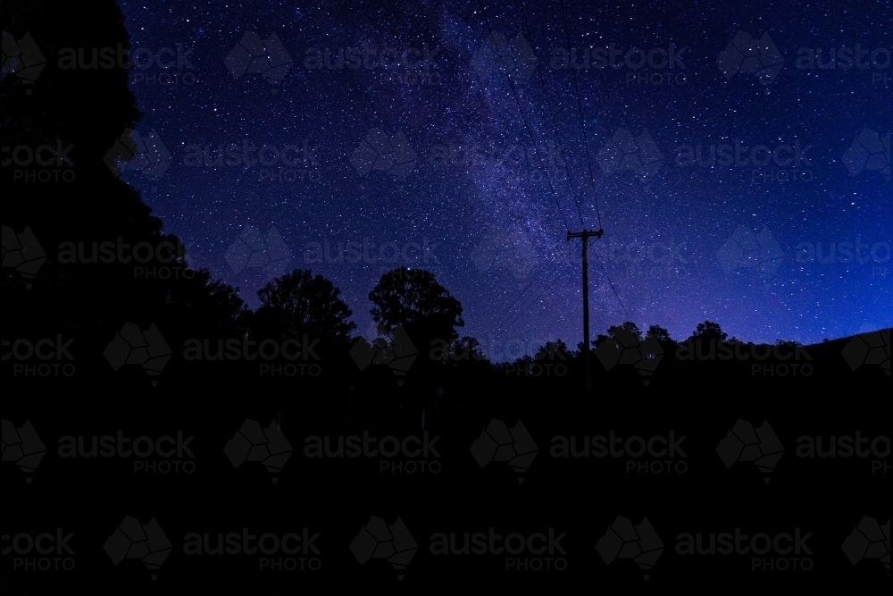 horizontal shot of some silhouette of trees, bushes and electric post of wires at dawn with stars - Australian Stock Image