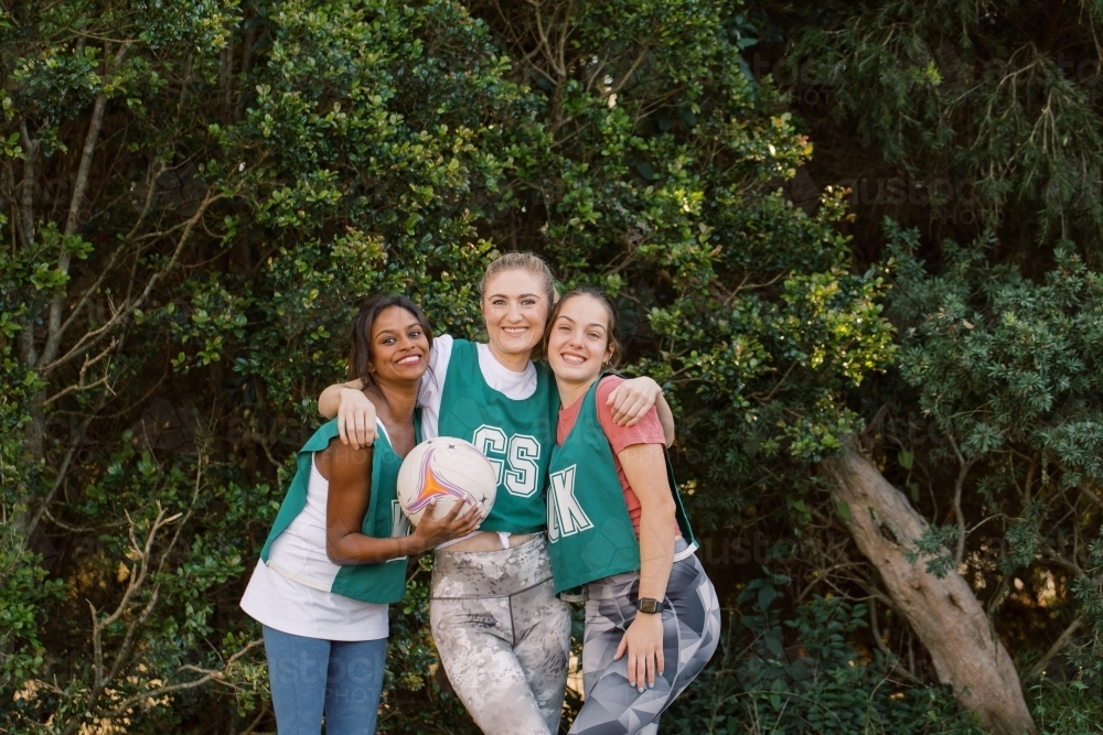 horizontal shot of three young smiling women posing outdoors with one holding a net ball - Australian Stock Image
