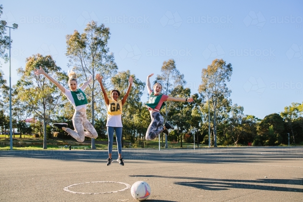 horizontal shot of three young women jumping in mid air with trees at the back and ball on ground - Australian Stock Image