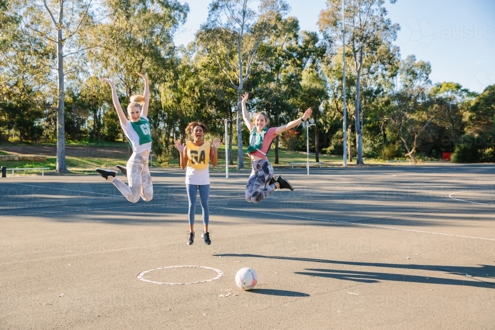 horizontal shot of three young women jumping in mid air with trees at the back and ball on ground - Australian Stock Image