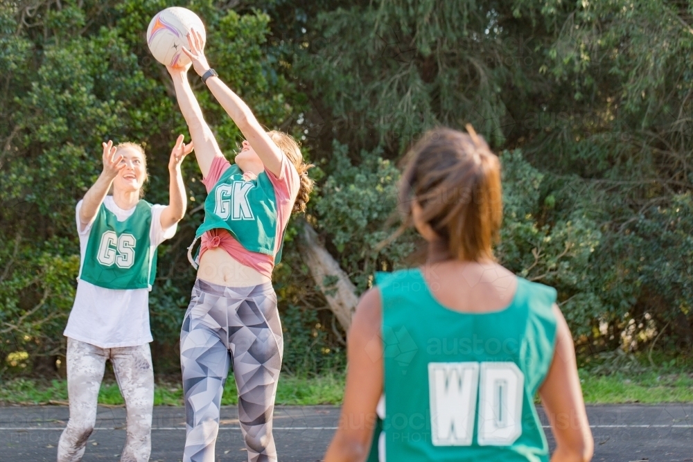 horizontal shot of three young women playing net ball on a sunny day with trees in the background - Australian Stock Image