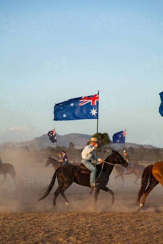 Horseman in akubra hat with Australian flag riding in dusty paddock - Australian Stock Image