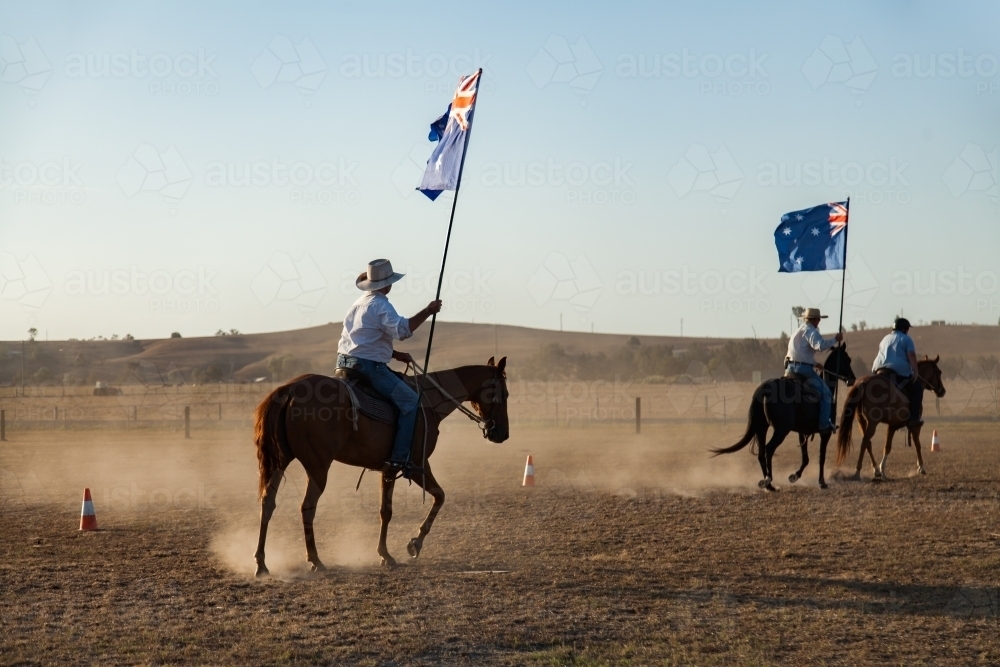 Horseman in akubra hat with Australian flag riding in dusty paddock - Australian Stock Image