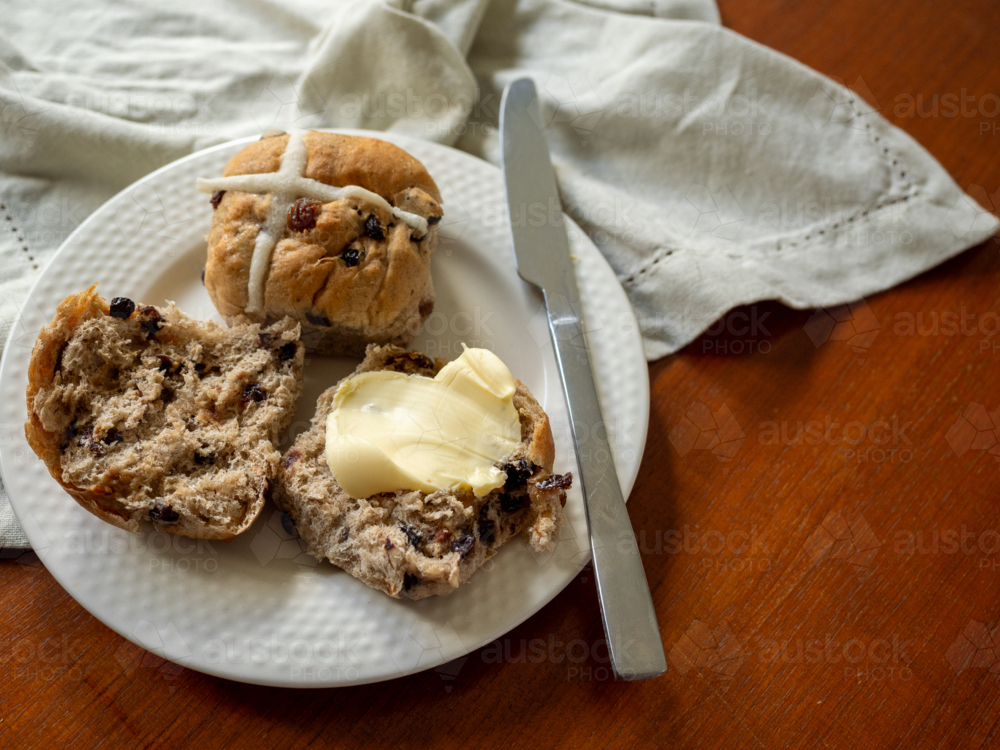 Hot cross buns with butter and knife on white plate, linen napkin and wooden tabletop - Australian Stock Image