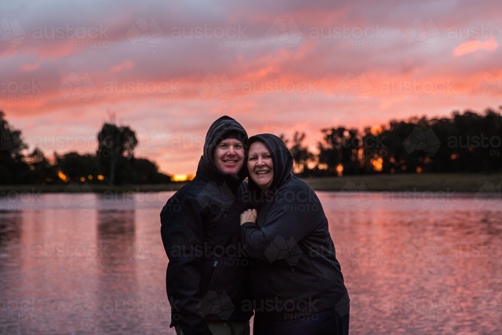 Husband and wife cuddling close together smiling happy with river water reflecting sunset sky - Australian Stock Image