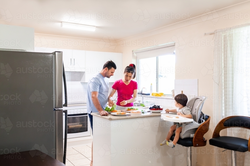 Husband and wife making Vietnamese fresh spring rolls in kitchen at home with toddler in chair - Australian Stock Image