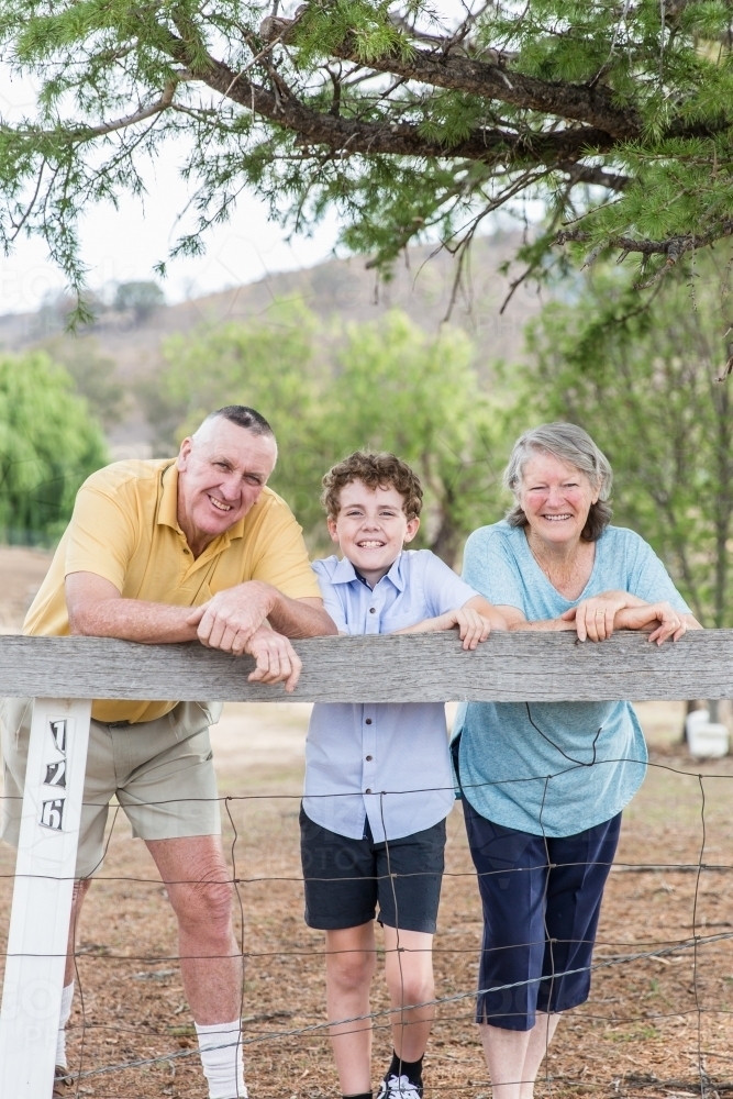 Husband and wife retired grandparents standing with grandson leaning on fence happy smiling - Australian Stock Image