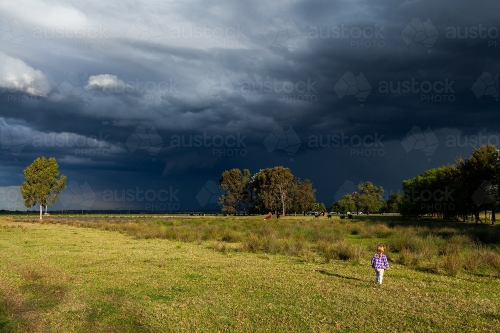 Incoming dark stormy rain clouds over sunlit rural farm paddock with country kid exploring - Australian Stock Image