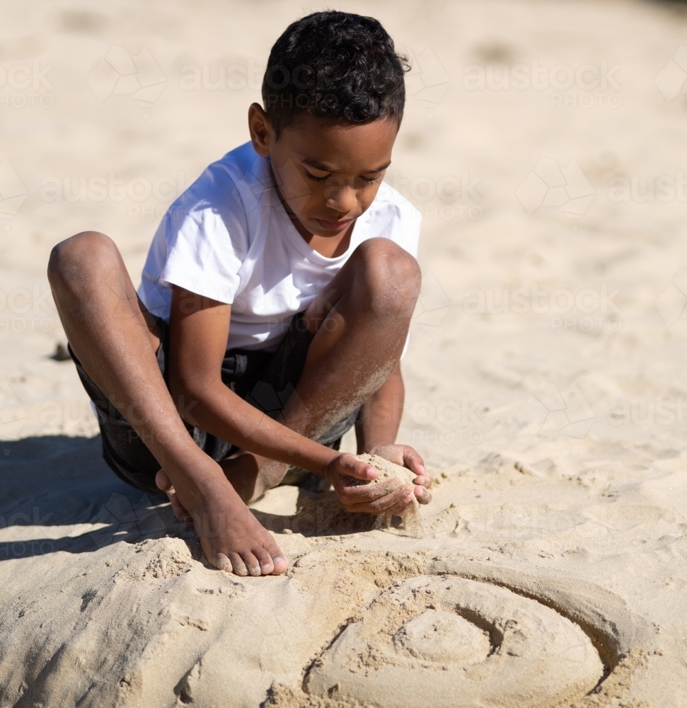 Indigenous boy creating design in wet sand - Australian Stock Image
