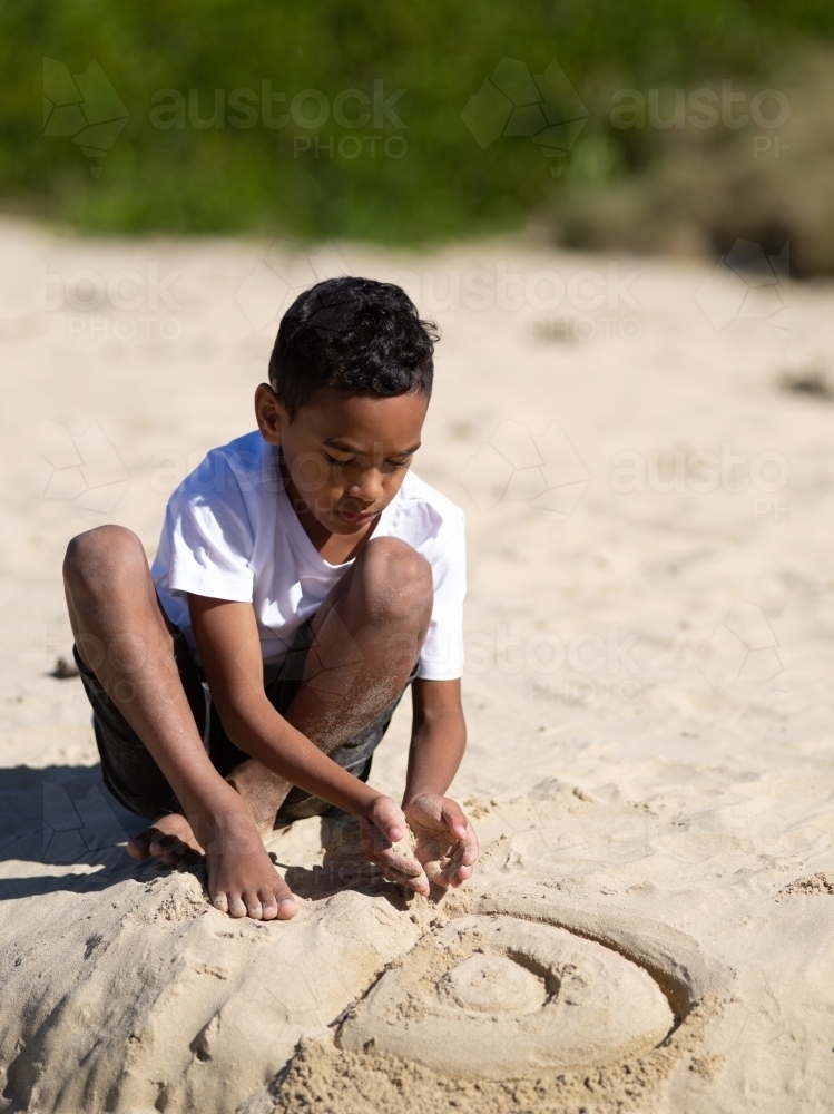 Indigenous boy creating design in wet sand - Australian Stock Image
