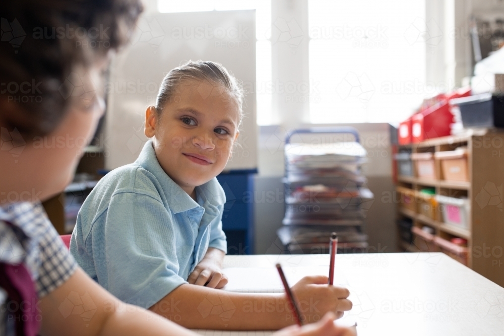 Indigenous girl smiling to her friend whilst writing in a primary school classroom - Australian Stock Image