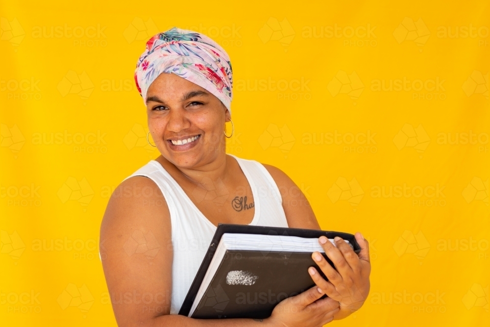 indigenous woman smiling and carrying books against yellow background - Australian Stock Image