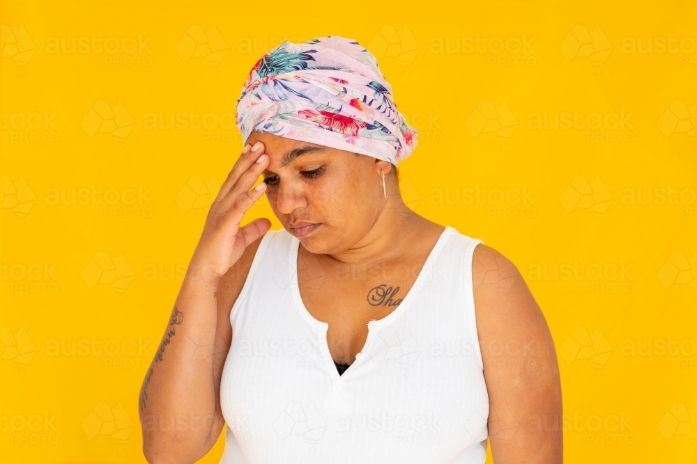 indigenous woman wearing head wrap with hand to forehead against yellow background - Australian Stock Image